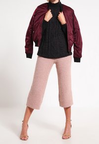 Giacca bomber bordeaux sopra un dolcevita in maglia nera a trama, abbinata a pantaloni cropped a gamba larga rosa chiaro e tacchi beige con fasce.