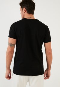 T-shirt nera a maniche corte con colletto rotondo, realizzata in morbido cotone. Presenta un tatuaggio visibile sul braccio sinistro.