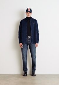 Camicia blu con bottoni sopra un dolcevita nero, abbinata a jeans blu e stivali neri. Un cappellino blu presenta un'accenno rosso e bianco.
