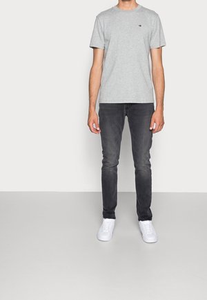 Mann trägt hellgraues kurzärmeliges T-Shirt, ausgewaschene schwarze Slim-Fit-Jeans und weiße Turnschuhe, steht vor einem einfarbigen hellen Hintergrund.