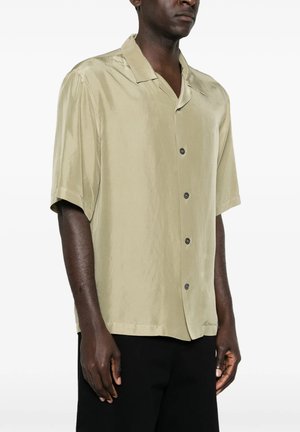Uomo che indossa una camicia beige larga a maniche corte abbottonata e pantaloni neri, in piedi davanti a uno sfondo bianco semplice.