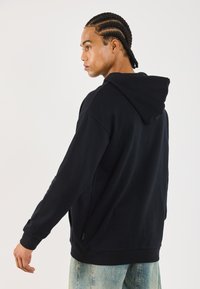 Svart hettegenser, mykt materiale, løs passform, kengurulomme, ribbestrikkede mansjetter, brukt med lyseblå jeans, som viser minimale sømmer og merkevare.
