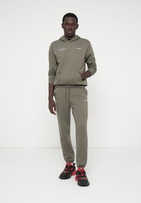 Hoodie verde-oliva e calças de moletom combinando, feitas de tecido suave. O hoodie tem um bolso canguru e detalhes em texto. Usado com ténis pretos e vermelhos.