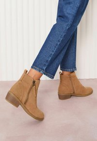 Világosbarna velúr bokacsizma oldalzippzárral, alacsony tömbsarokkal és texturált talppal, kék rövid szárú farmernadrággal párosítva.