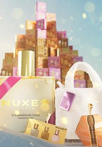 Kalendarz adwentowy NUXE w złotym i różowym opakowaniu, w towarzystwie białej, puszystej torby, małych próbek produktów oraz buteleczki w kolorze bursztynu z złotymi akcentami.