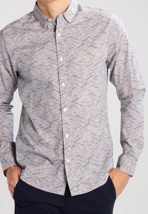 Muž nosící košili s dlouhým rukávem zapnutou na knoflíky s vířivým vlnovým vzorem v tmavě modré a bílé barvě, rukama v kapsách tmavě modrých kalhot.