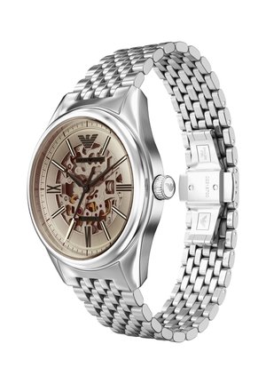 Reloj de acero inoxidable con correa de malla plateada, esfera esqueletizada beige, números romanos y caja pulida. Cuenta con logotipo grabado en el lateral.