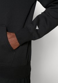 Sudadera negra con bolsillo canguro, puños y dobladillo acanalados, y un pequeño logo blanco en la manga. Textura de tela suave.