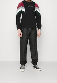 Persona vistiendo pantalones de chándal negros, zapatillas deportivas bajas negras y blancas, y una sudadera negra con rayas de colores y el texto "KAOTIKO".