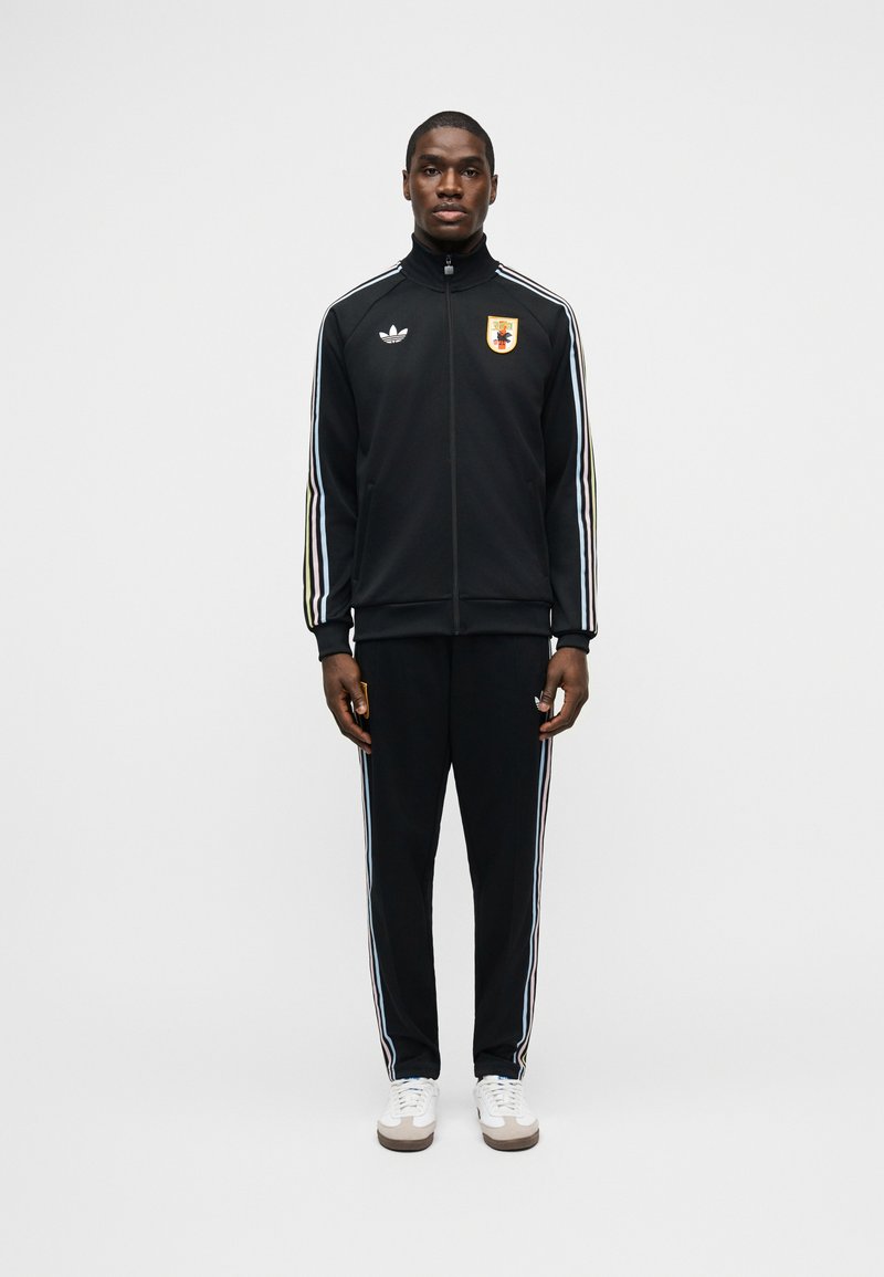 Mand står iført sort Adidas træningsdragt med hvide striber og broderet emblem, kombineret med hvide sneakers, foran ensfarvet baggrund.