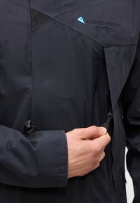 Χέρι τραβά το φερμουάρ σε μαύρο μπουφάν Klättermusen Allgrön 2.0, αποκαλύπτοντας την υφή του υφάσματος και τη λεπτομέρεια του λογότυπου.