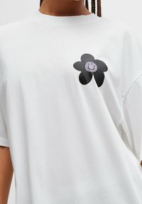 Biały bawełniany t-shirt z czarnym kwiatowym wzorem, którego środek tworzy uśmiechnięta twarz. Krótkie rękawy i luźny krój.