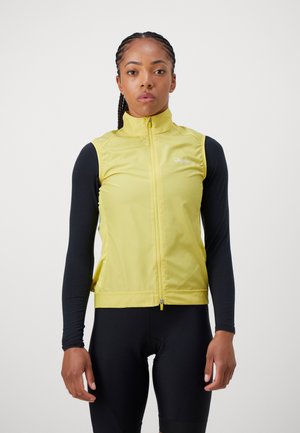 Yellow sleeveless cycling vest with high collar, zip closure, and Rapha logo. Worn over a black long-sleeve top and black shorts.