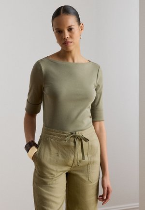 Femme aux cheveux attachés portant un haut vert olive à manches mi-longues et un pantalon taille haute avec cordon, debout avec une main dans la poche.