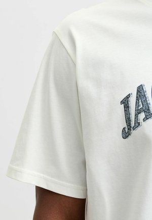 T-shirt à manches courtes crème clair avec un imprimé graphique noir portant le mot « JACK » sur le devant, fabriqué en coton doux et lisse.