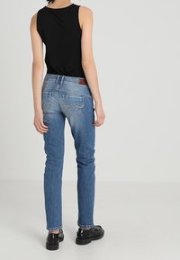 Jeans de mezclilla azul con corte slim, textura desvaída y diseño clásico de cinco bolsillos. Combinados con una blusa sin mangas negra y zapatos negros.
