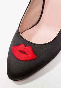 Escarpin en satin noir à bout pointu, ornée de lèvres rouges brodées sur la tige. Texture lisse avec des détails de couture minimaux.