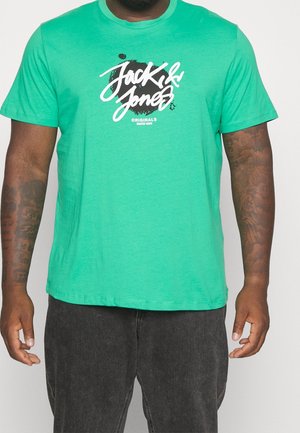 Homme portant un t-shirt vert vif Jack & Jones avec un texte graphique blanc, associé à un jean noir, montrant des tatouages sur les deux avant-bras.