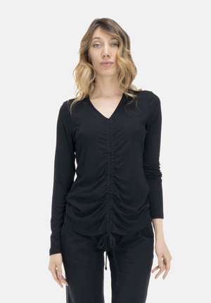 Femme aux cheveux blonds ondulés portant un haut noir à manches longues froncé avec un col en V et un pantalon noir, debout devant un fond clair uni.