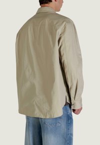 Camicia beige con bottoni, maniche lunghe, vestibilità comoda, spacchi laterali e bottoni color argento. Indossata sopra jeans larghi azzurri.