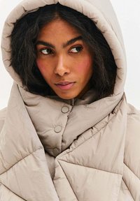 Béžová prošívaná bunda s velkou kapucí, zdobená prošitým geometrickým vzorem a zapínáním na knoflíky. Měkký, texturovaný materiál.