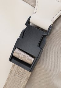Čierna plastová pracka s béžovým páskom, pripevnená k svetlému, hladkému materiálu. Má viditeľné švy a textúrovaný povrch.