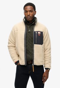 Beige Fleecejacke mit einer blauen Tasche, Reißverschluss und hohem Kragen. Verfügt über einen strukturierten Stoff, Brustpatch und kontrastierende Ärmel-Details.