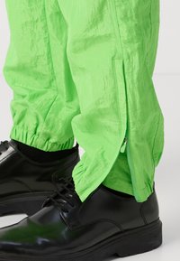 Neon zelene, lagane hlače s bočnim zipovima i elastičnim manžetama, uparene s sjajnim crnim cipelama.