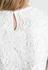 Vit spetsblus med blommönster, texturerad tyg och en nyckelhålsdetalj vid baksidan av halsringningen. Kantad med en delikat scalloped trim.