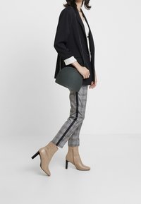 Bottines beiges à talons avec des pointes aiguës, associées à un pantalon à carreaux gris avec des rayures noires et un blazer noir. Accentué par un sac vert.