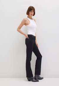 Donna con i capelli corti castani che indossa una canotta bianca senza maniche, pantaloni aderenti neri e stivaletti neri, in piedi contro uno sfondo bianco semplice.