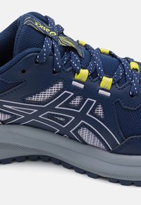 Blauwe en grijze ASICS hardloopschoen met mesh-panelen, gele vetersluitingen en gespikkelde blauwe veters op een witte achtergrond.