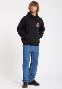 Sort hætte sweatshirt med frontlomme, der har et flerfarvet broderet design. Båret med blå jeans og sorte sko.
