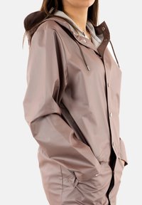 Giacca impermeabile marrone con cappuccio, dotata di chiusura a bottoni, cordini e due tasche laterali. Texture liscia e lucida.