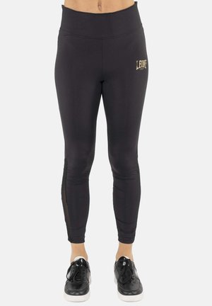 Femme portant des leggings noirs taille haute avec un petit logo doré "LEONE 1947" sur la partie supérieure de la cuisse gauche et des baskets noires à semelles blanches.