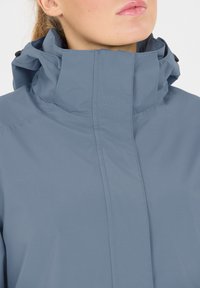 Ljusblå vattentät jacka med hög krage, slät textur och synlig sömnad. Har knappar för justering av kragen och dragkedja.