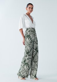 Bela srajca s kratkimi rokavi in gumbi, kombinirana z zelenimi širokimi hlačami z belim vzorcem palminega lista ter sandali z visokimi petami.