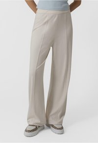 Pantalons large beige clair avec des coutures verticales, texture lisse, et taille haute, assortis à des baskets à motifs.