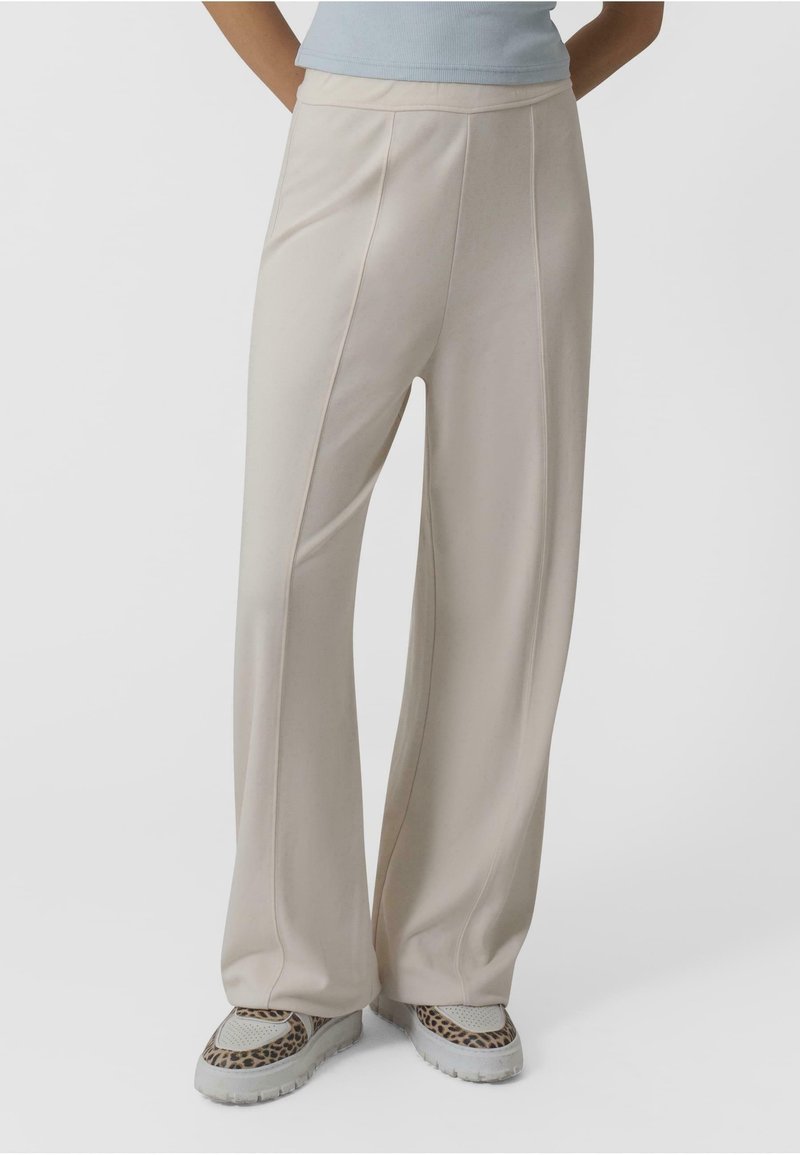 Pantalons large beige clair avec des coutures verticales, texture lisse, et taille haute, assortis à des baskets à motifs.