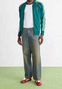 Tealfärgad zip-up jacka med gula ränder, i kombination med bleka blå vida jeans och röda skor med vita ränder. Vit linne under.