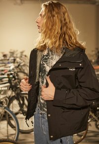 Sort vandtæt jakke med justerbar hætte og foran knapper, båret over en mønstret skjorte. Baggrunden indeholder cykler.