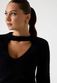 Suéter negro de pelaje suave con un escote en V profundo y un diseño recortado en el cuello. La modelo lleva pendientes de aro dorados, mostrando una tela texturizada.