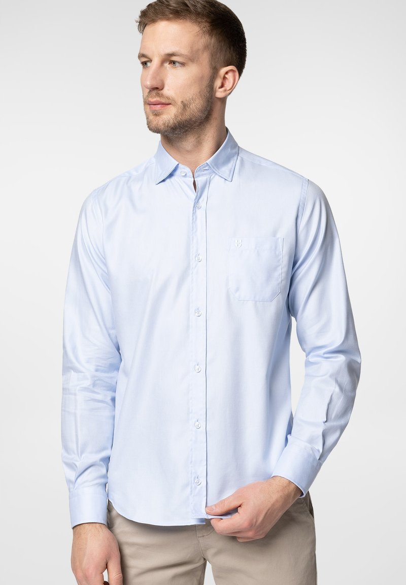 Jasnoniebieska, długa koszula zapinana na guziki z usztywnianym kołnierzykiem, jedną kieszenią na piersi i gładką fakturą materiału.