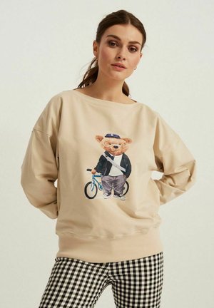 Beżowy, oversize'owy sweter z grafiką misia w kurtce, trzymającego niebieski rower. Czarno-białe spodenki w kratę. Miękka tkanina, luźny krój.