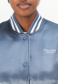 Jachetă bomber din satin albastru, cu guler în dungi, închidere cu nasturi și logo-ul brodat alb "tommy jeans" pe piept.