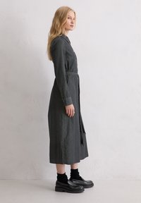 Długa sukienka zapinana na guziki w ciemnoszarym materiale, z kołnierzykiem, długimi rękawami, paskiem w talii i długością do połowy łydki. Noszona z czarnymi butami i skarpetkami.