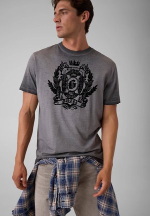 Ung mand iført en grå t-shirt med et sort udsmykket våbenskjold, lyse bukser og en blå ternet skjorte bundet om livet, kigger til siden.