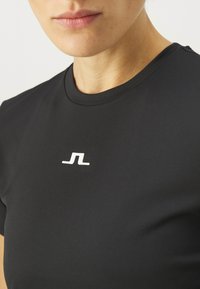 Juodas sportinis marškinėliai su trumpo rankovėmis, prigludusio dizaino, pagaminti iš lygaus audinio. Yra mažas baltas logotipas krūtinės srityje.