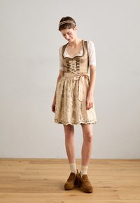La robe présente un corsage ajusté en tissu beige avec un devant lacé et de courtes manches en dentelle, associé à une jupe fluide, brodée et ornée d'un ruban en satin.