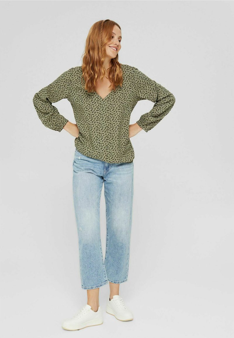 Olijfgroene blouse met lange mouwen, een diepe V-hals en een klein zwart bloemenpatroon, gecombineerd met lichtblauwe cropped jeans en witte sneakers.